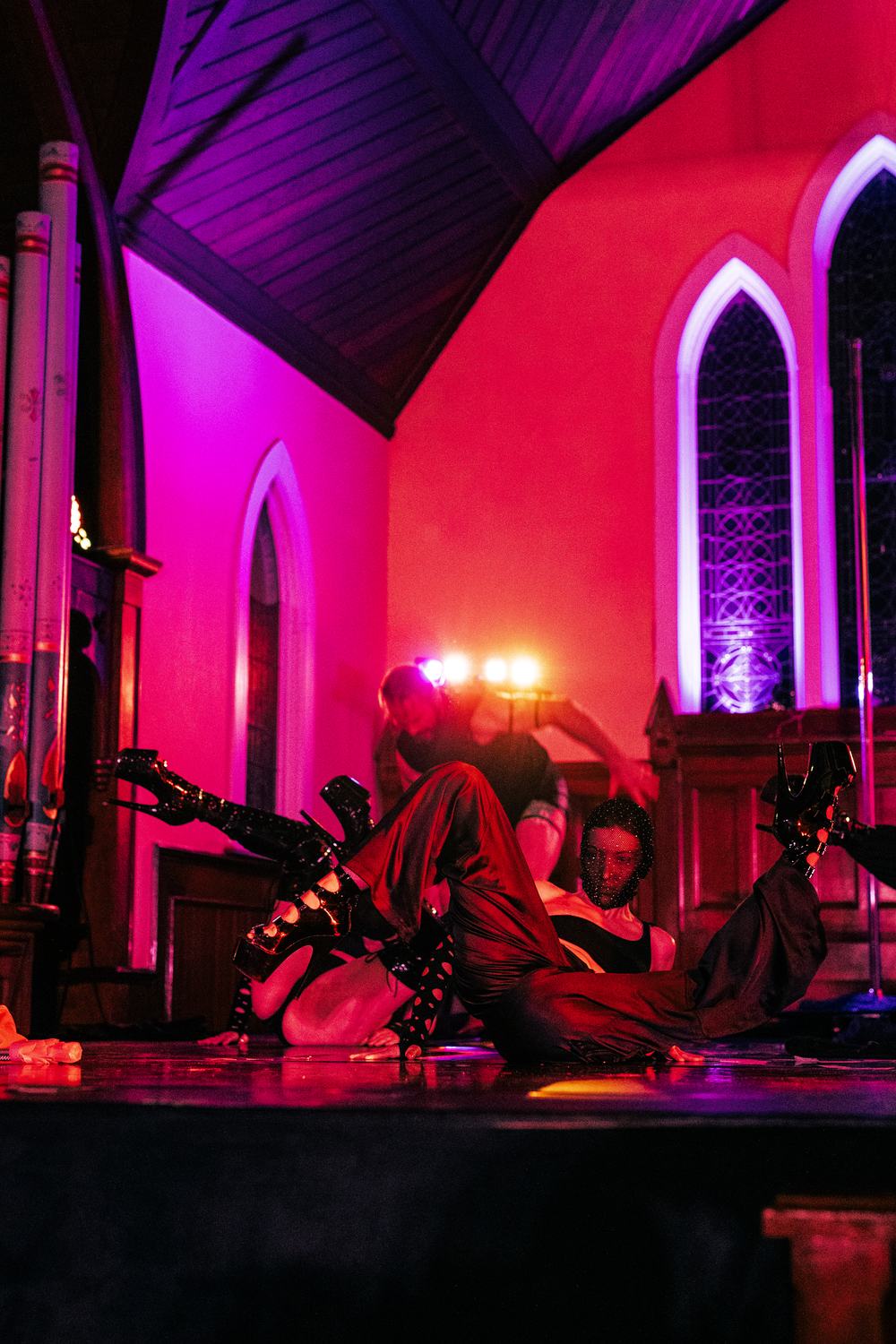 Two dancers in platform boots on the floor of a church venue under magenta and red stage light, gothic arched windows and organ pipes behind them