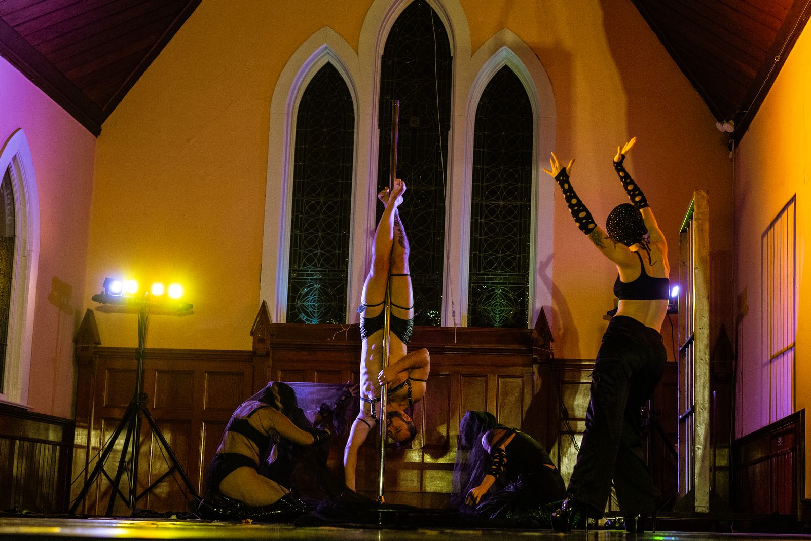 A PoleCoven performer inverted on the pole in the church venue, warm gold light, dancers gathered at the base.
