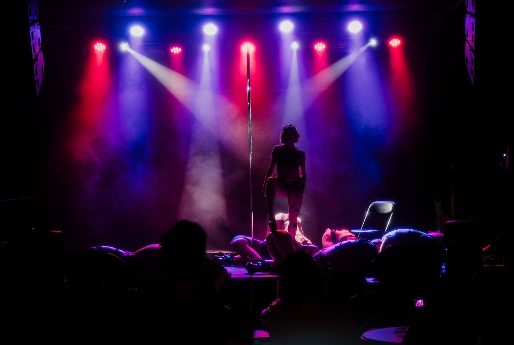 A performer in a crown on a smoke-filled stage under red and purple lights, audience silhouetted in front